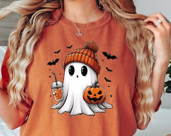 May include: A woman wearing an orange t-shirt with a cute cartoon ghost holding a pumpkin and a cup of coffee. The ghost is wearing a brown and orange hat. The shirt has a Halloween theme.