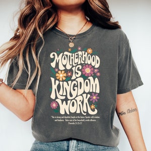 May include: A dark gray t-shirt with a floral design and the text "MOTHERHOOD IS KINGDOM WORK" in a retro font. The text is surrounded by colorful flowers. The shirt also includes a quote from Proverbs 31:25-27.