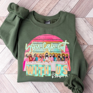 May include: Olive green sweatshirt with a graphic featuring the text "you can sit with us" above a depiction of the Last Supper. The design includes a pink and orange sunset and the text "Romans 15".