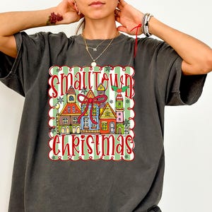 May include: Dark grey t-shirt with a colourful graphic that reads "Smalltown Christmas" and depicts houses. The design is framed by a green and white striped border with red accents, creating a festive holiday theme.