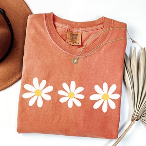 May include: A coral colored t-shirt with three white daisy flowers printed on the front. The shirt has a crew neck and is made of a soft, comfortable fabric. The shirt is folded and laid flat on a white surface.