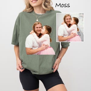 May include: A moss green t-shirt featuring a printed image of a man and a child. The image shows the man embracing the child, who is wearing a pink tutu. The word "Moss" is printed in black at the top right.