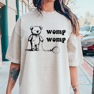 May include: A woman wearing a beige oversized t-shirt with a black and white illustration of a teddy bear holding a balloon and the text "womp womp". She is wearing white sneakers.