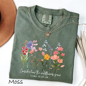 May include: Moss green t-shirt with a floral design and the text "Consider how the wildflowers grow" from Luke 12:27-28. The shirt has a crew neck and short sleeves. A silver necklace with a small pendant is on the shirt.