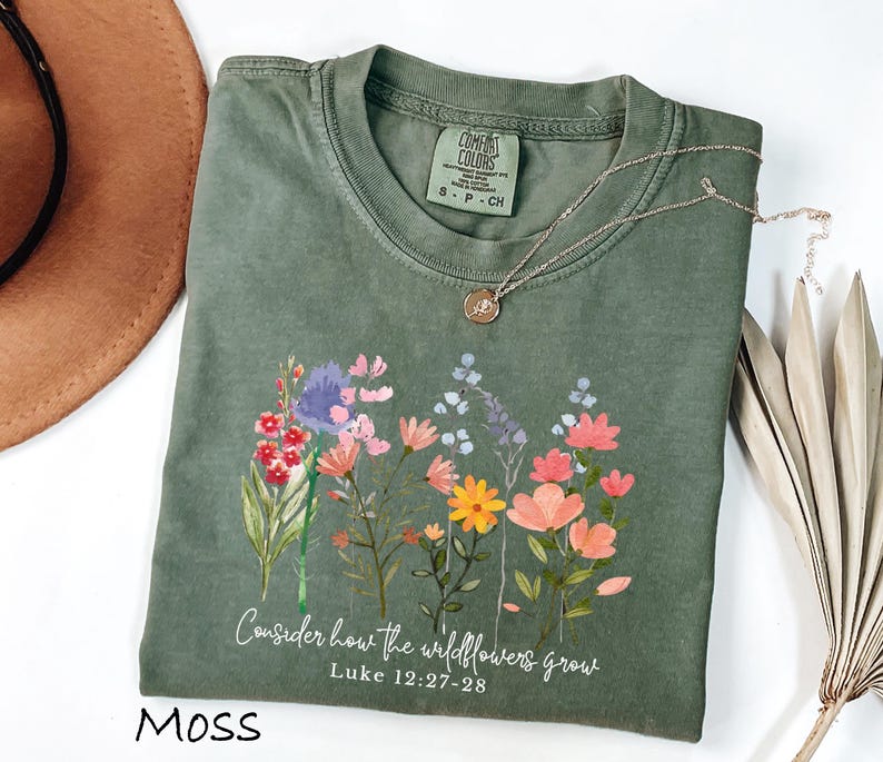 May include: Moss green t-shirt with a floral design and the text "Consider how the wildflowers grow" from Luke 12:27-28. The shirt has a crew neck and short sleeves. A silver necklace with a small pendant is on the shirt.