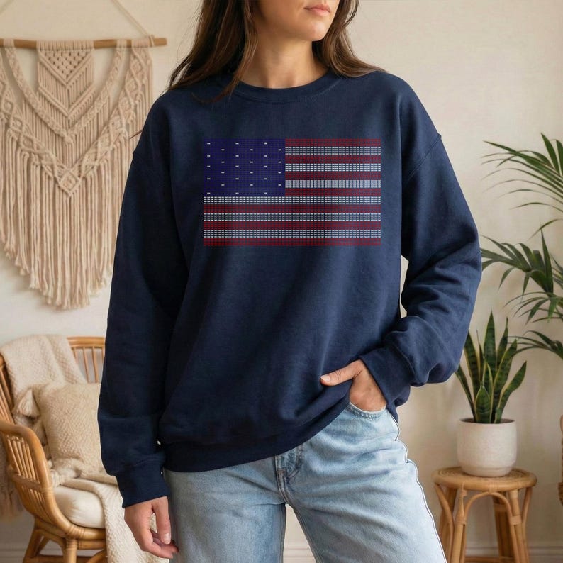 A woman wearing a navy blue sweatshirt with an American flag design stands in a room with a woven wall hanging and potted plants.