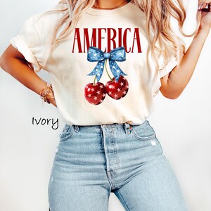 May include: A white t-shirt with the word "AMERICA" printed in red letters. A blue bow with white stars is above the word, and two red cherries with white stars are below the bow.