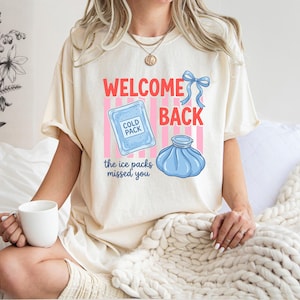 May include: An ivory t-shirt featuring a graphic with the text "WELCOME BACK" in red, a blue bow, a light blue cold pack, and a blue ice bag. Below the graphics, the text "the ice packs missed you" is printed in blue, set against pink stripes.