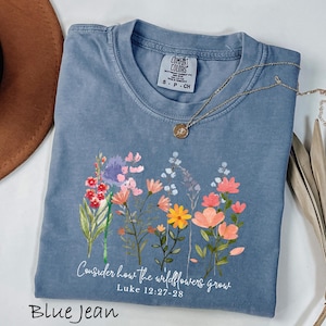 May include: Blue jean colored t-shirt with a floral design and the text "Consider how the wildflowers grow" and "Luke 12:27-28". The shirt has a crew neck and short sleeves. A gold necklace with a pendant is draped across the shirt.