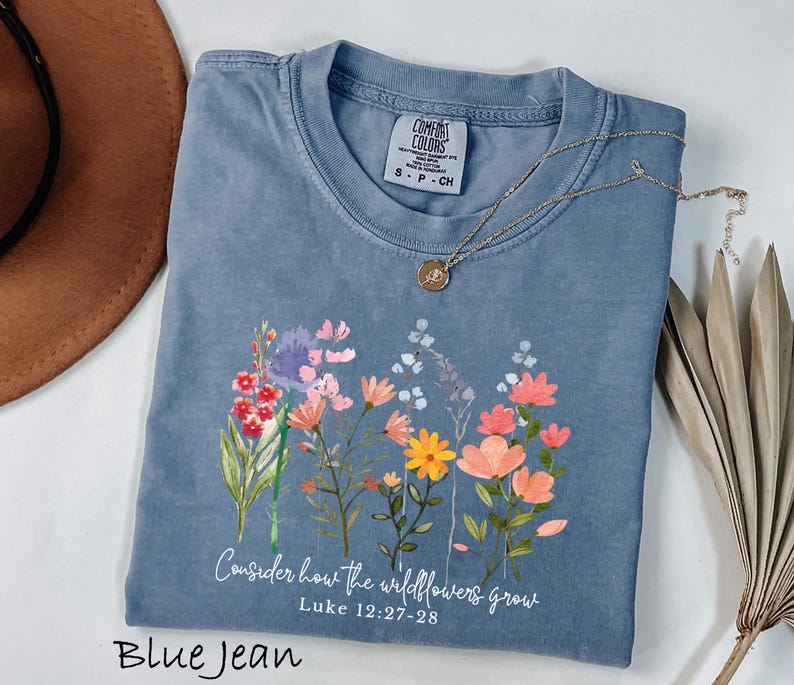 May include: Blue jean colored t-shirt with a floral design and the text "Consider how the wildflowers grow" and "Luke 12:27-28". The shirt has a crew neck and short sleeves. A gold necklace with a pendant is draped across the shirt.