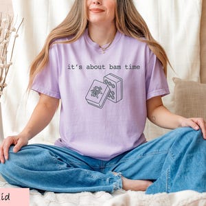 A woman wearing a purple t-shirt with a graphic design sits cross-legged on a bed, with a vase of flowers and a beige pillow nearby.
