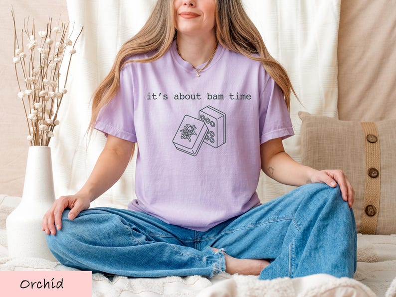A woman wearing a purple t-shirt with a graphic design sits cross-legged on a bed, with a vase of flowers and a beige pillow nearby.