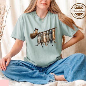 A woman wearing a green t-shirt with a graphic of cats sitting on a broom, sitting on a bed with a vase of flowers and a pillow nearby.