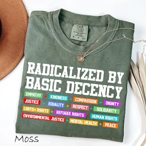 May include: A moss green t-shirt with the text "RADICALIZED BY BASIC DECENCY" in white block letters. Below the text are colorful rectangles with words like "EMPATHY", "JUSTICE", and "EQUALITY". A gold necklace with a pendant is draped across the shirt.
