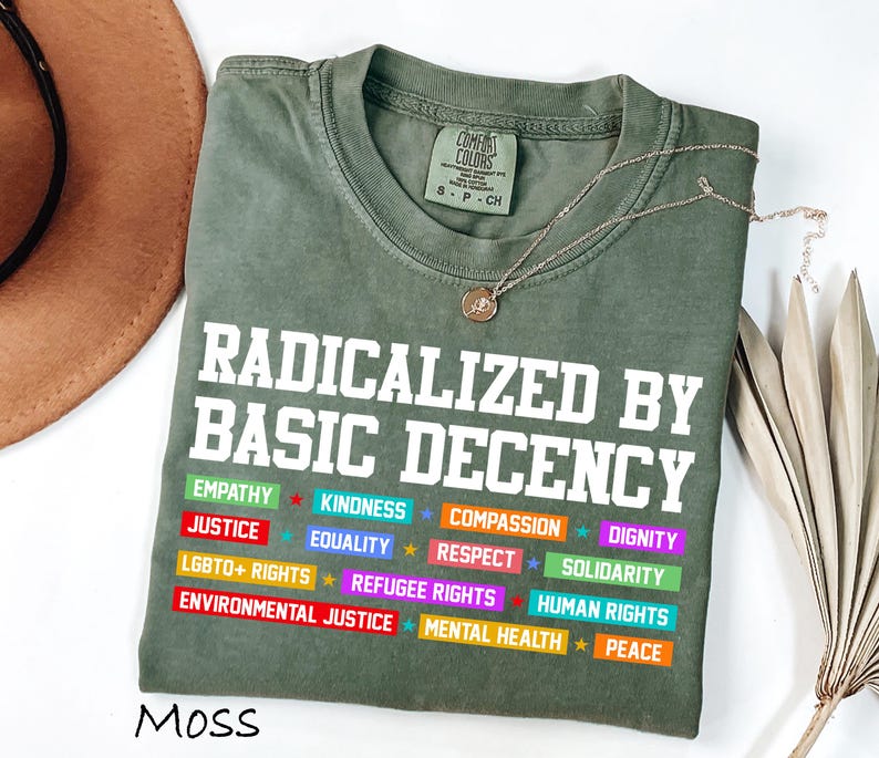 May include: A moss green t-shirt with the text "RADICALIZED BY BASIC DECENCY" in white block letters. Below the text are colorful rectangles with words like "EMPATHY", "JUSTICE", and "EQUALITY". A gold necklace with a pendant is draped across the shirt.