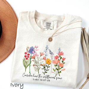 May include: Ivory-colored Comfort Colors t-shirt with a watercolor floral design. The design includes various wildflowers in shades of pink, blue, yellow, and red. The text "Consider how the wildflowers grow" is printed below the flowers.