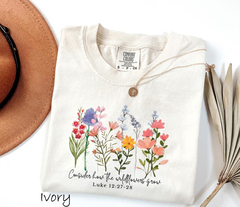 May include: Ivory-colored Comfort Colors t-shirt with a watercolor floral design. The design includes various wildflowers in shades of pink, blue, yellow, and red. The text "Consider how the wildflowers grow" is printed below the flowers.