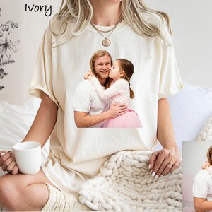 May include: Ivory-colored t-shirt featuring a printed image of a man and a child. The child is kissing the man on the cheek. The t-shirt is worn by a person holding a white mug. The word "ivory" is printed at the top.