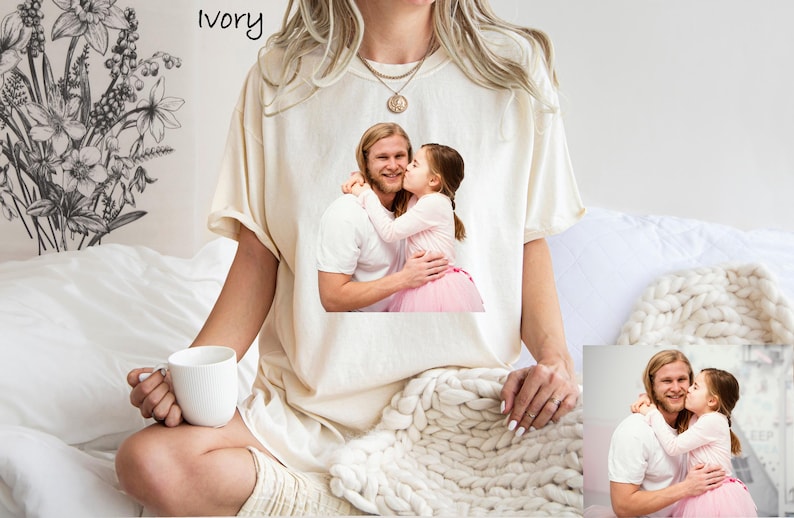 May include: Ivory-colored t-shirt featuring a printed image of a man and a child. The child is kissing the man on the cheek. The t-shirt is worn by a person holding a white mug. The word "ivory" is printed at the top.
