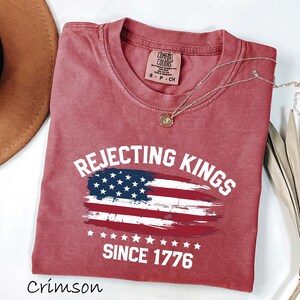 May include: Crimson-colored t-shirt with the text "Rejecting Kings Since 1776" and an American flag design. The shirt has a crew neck and short sleeves. A gold necklace is draped across the shirt.