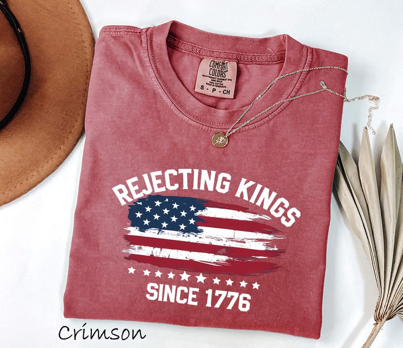 May include: Crimson-colored t-shirt with the text "Rejecting Kings Since 1776" and an American flag design. The shirt has a crew neck and short sleeves. A gold necklace is draped across the shirt.
