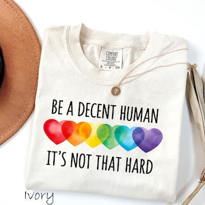 May include: Ivory-colored t-shirt with the text "BE A DECENT HUMAN IT'S NOT THAT HARD" in black. The design includes a row of rainbow-colored hearts. A gold necklace and a brown hat are also visible.
