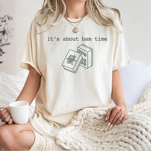 A woman wearing a white t-shirt with a graphic design and holding a white mug is sitting on a bed with a white blanket.