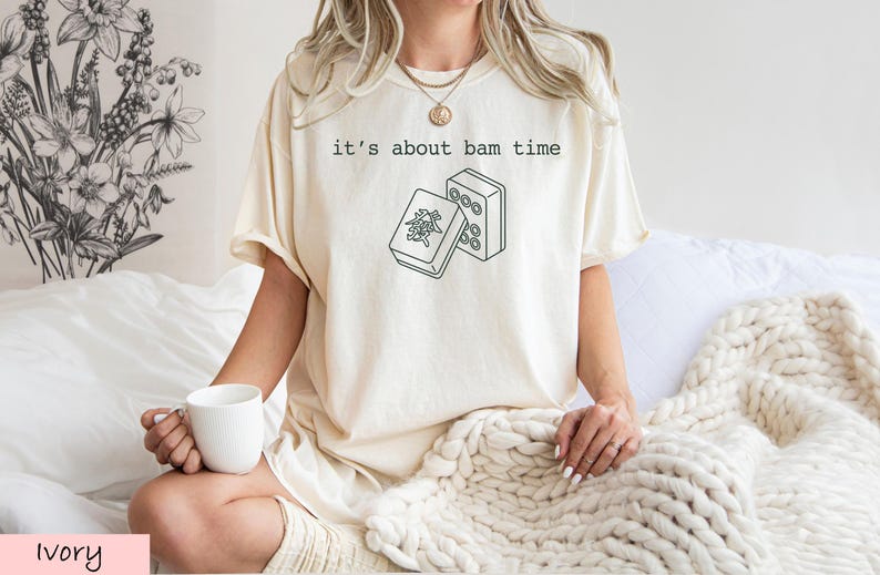 A woman wearing a white t-shirt with a graphic design and holding a white mug is sitting on a bed with a white blanket.