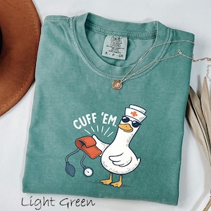May include: Light green t-shirt with a graphic of a goose wearing sunglasses and a nurse's hat, holding a blood pressure cuff. The text "CUFF 'EM" is above the goose. A gold necklace is draped on the shirt.
