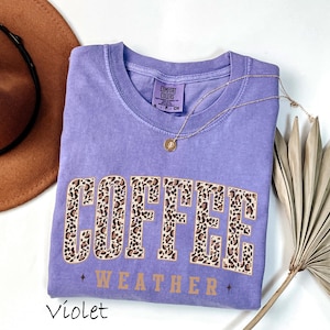 May include: A violet Comfort Colors t-shirt with the words "COFFEE WEATHER" in a leopard print design. A gold necklace and a brown hat are next to the shirt. The shirt is a size small.