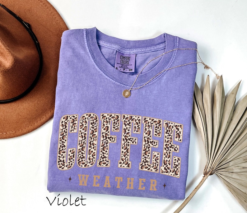 May include: A violet Comfort Colors t-shirt with the words "COFFEE WEATHER" in a leopard print design. A gold necklace and a brown hat are next to the shirt. The shirt is a size small.