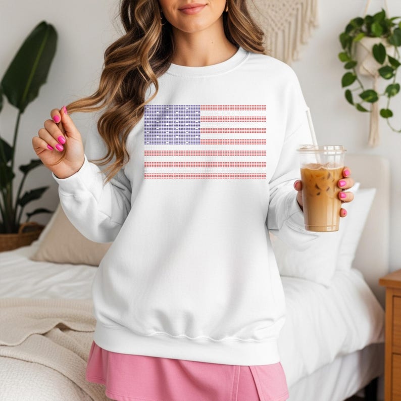 A woman wearing a white sweatshirt with an American flag design is holding a drink in her hand.