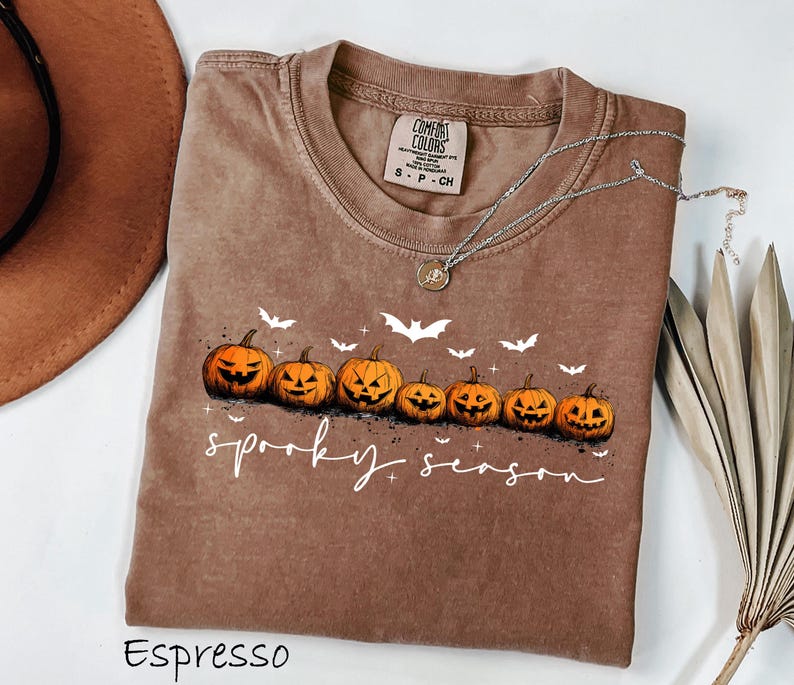 May include: Espresso-colored t-shirt with a crew neck, featuring a row of carved orange pumpkins and white bats. The words "spooky season" are written in white script. A silver necklace with a pendant is draped on the shirt.
