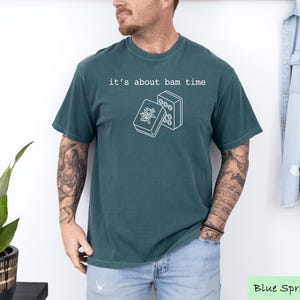 A man wearing a green t-shirt with the text "it's about bam time" printed on it, standing in front of a plant and a denim jacket.