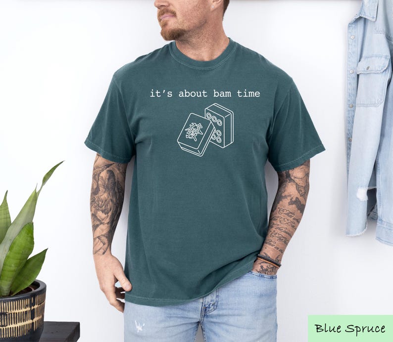 A man wearing a green t-shirt with the text "it's about bam time" printed on it, standing in front of a plant and a denim jacket.