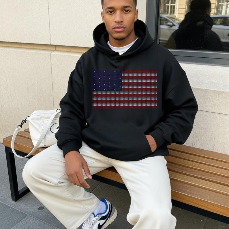 A young man wearing a black hoodie with an American flag design is sitting on a wooden bench.