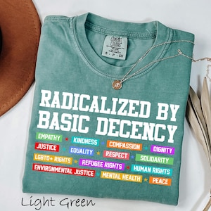 May include: Light green t-shirt with the text "RADICALIZED BY BASIC DECENCY" in white block letters. Below the text are colorful words like "EMPATHY", "JUSTICE", and "EQUALITY". A gold necklace with a pendant is draped across the shirt.