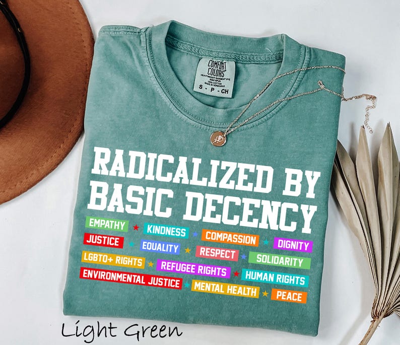 May include: Light green t-shirt with the text "RADICALIZED BY BASIC DECENCY" in white block letters. Below the text are colorful words like "EMPATHY", "JUSTICE", and "EQUALITY". A gold necklace with a pendant is draped across the shirt.