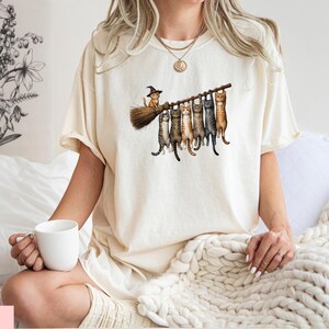 A woman wearing a white t-shirt with a graphic of cats on a broom is sitting on a bed, holding a white mug.