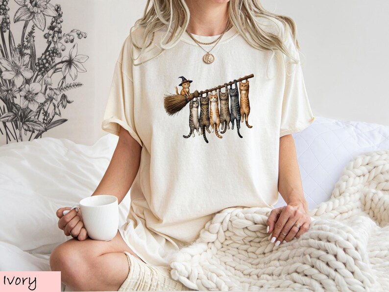 A woman wearing a white t-shirt with a graphic of cats on a broom is sitting on a bed, holding a white mug.