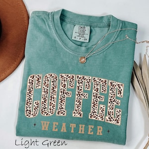 May include: A light green t-shirt featuring the words "COFFEE WEATHER" in a leopard print design. The shirt has a crew neck and short sleeves. A gold necklace with a pendant is draped across the shirt.