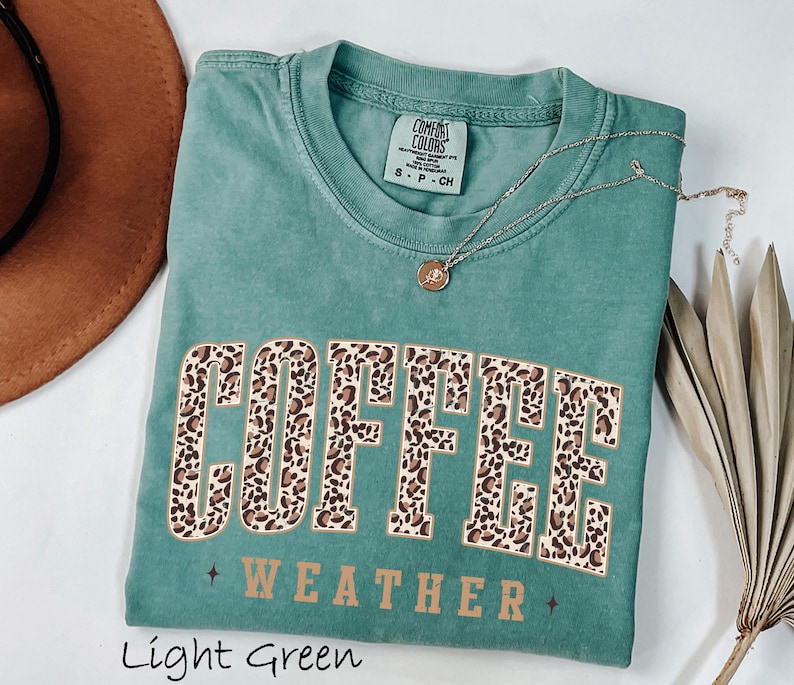 May include: A light green t-shirt featuring the words "COFFEE WEATHER" in a leopard print design. The shirt has a crew neck and short sleeves. A gold necklace with a pendant is draped across the shirt.