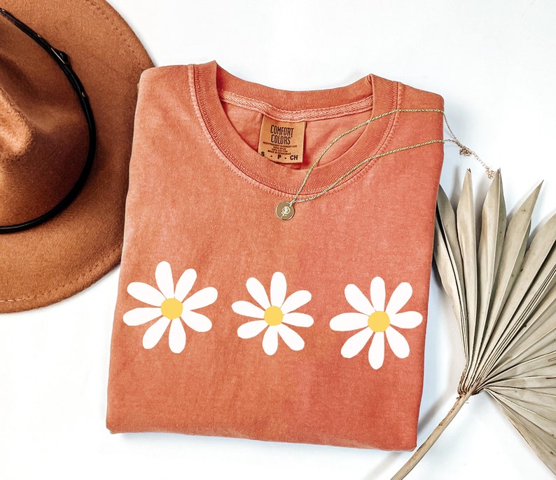 May include: A coral colored t-shirt with three white daisy flowers printed on the front. The shirt has a crew neck and is made of a soft, comfortable fabric. The shirt is folded and laid flat on a white surface.