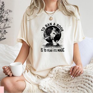 A woman wearing a white t-shirt with a graphic design and text, sitting on a bed with a white blanket.