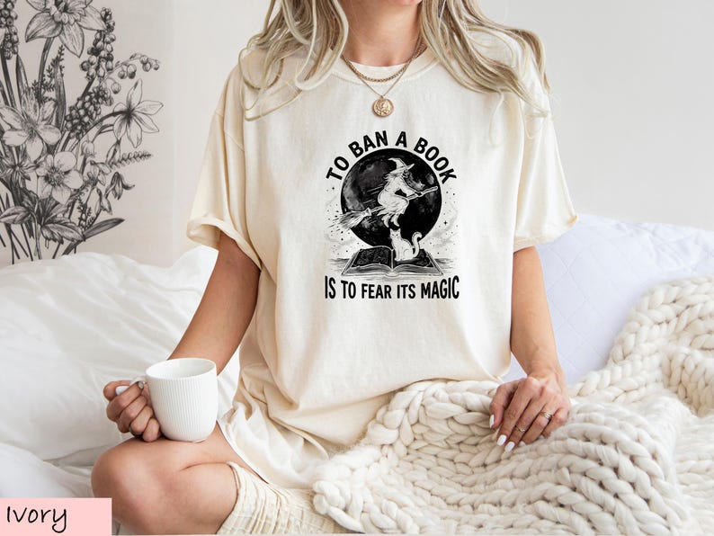 A woman wearing a white t-shirt with a graphic design and text, sitting on a bed with a white blanket.