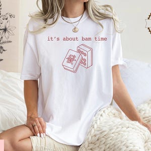 A woman wearing a white t-shirt with a red graphic and text printed on it, sitting on a bed with a white blanket.