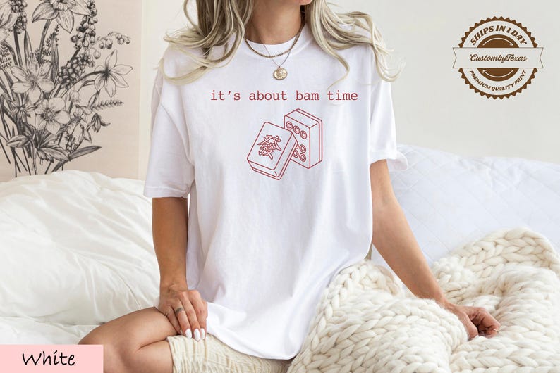 A woman wearing a white t-shirt with a red graphic and text printed on it, sitting on a bed with a white blanket.