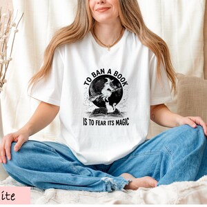A woman wearing a white t-shirt with a graphic design and text, sitting cross-legged on a bed with a vase of flowers in the background.