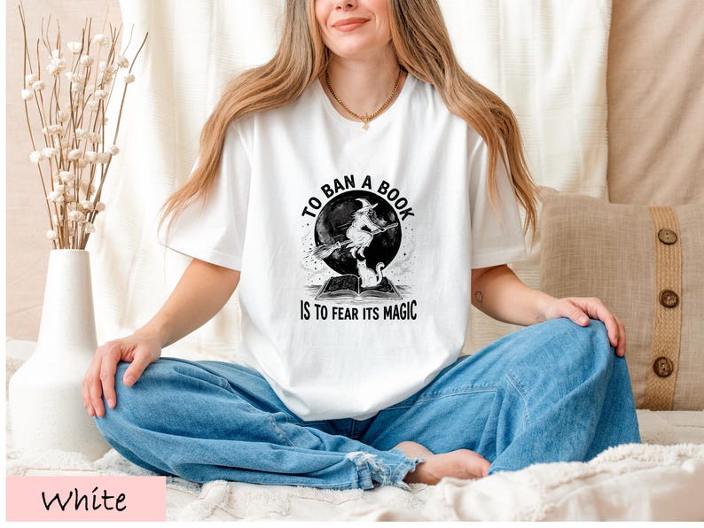 A woman wearing a white t-shirt with a graphic design and text, sitting cross-legged on a bed with a vase of flowers in the background.