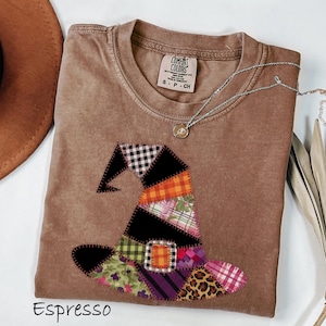 May include: Espresso-colored t-shirt with a patchwork witch hat design. The hat features various patterns, including plaid, leopard print, and solid colors. A silver necklace with a pendant rests on the shirt. The word "Espresso" is printed at the bottom.